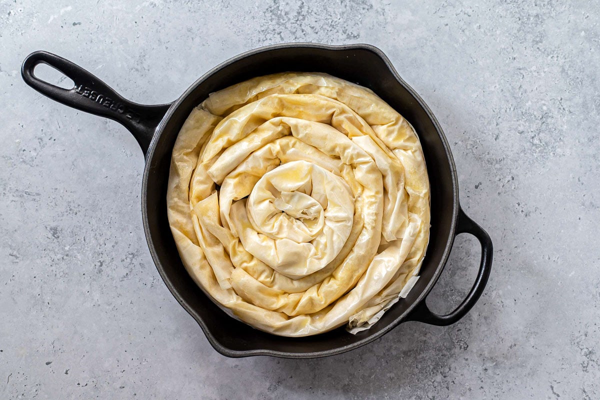 phyllo sheets filled with cheese rolled into spiral and placed in round black baking pan.