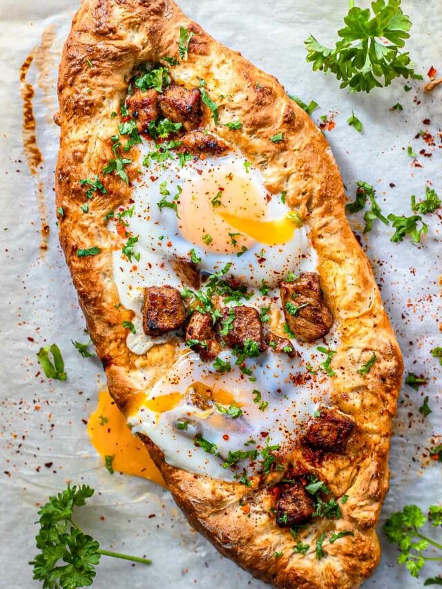 oval shaped pizza on parchment paper with pork chunks and egg on top.