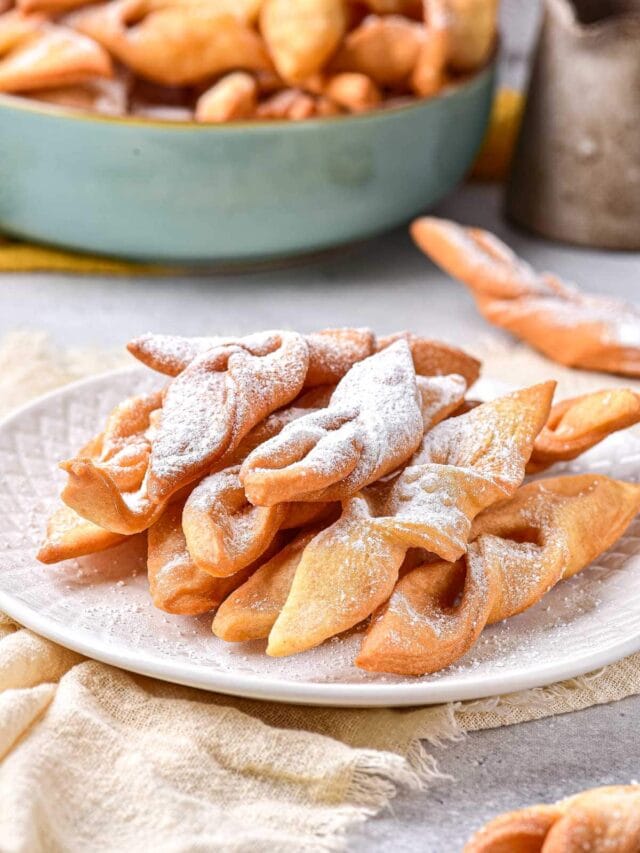 crispy fried dough cookies on plate covered in powdered sugar with more behind.