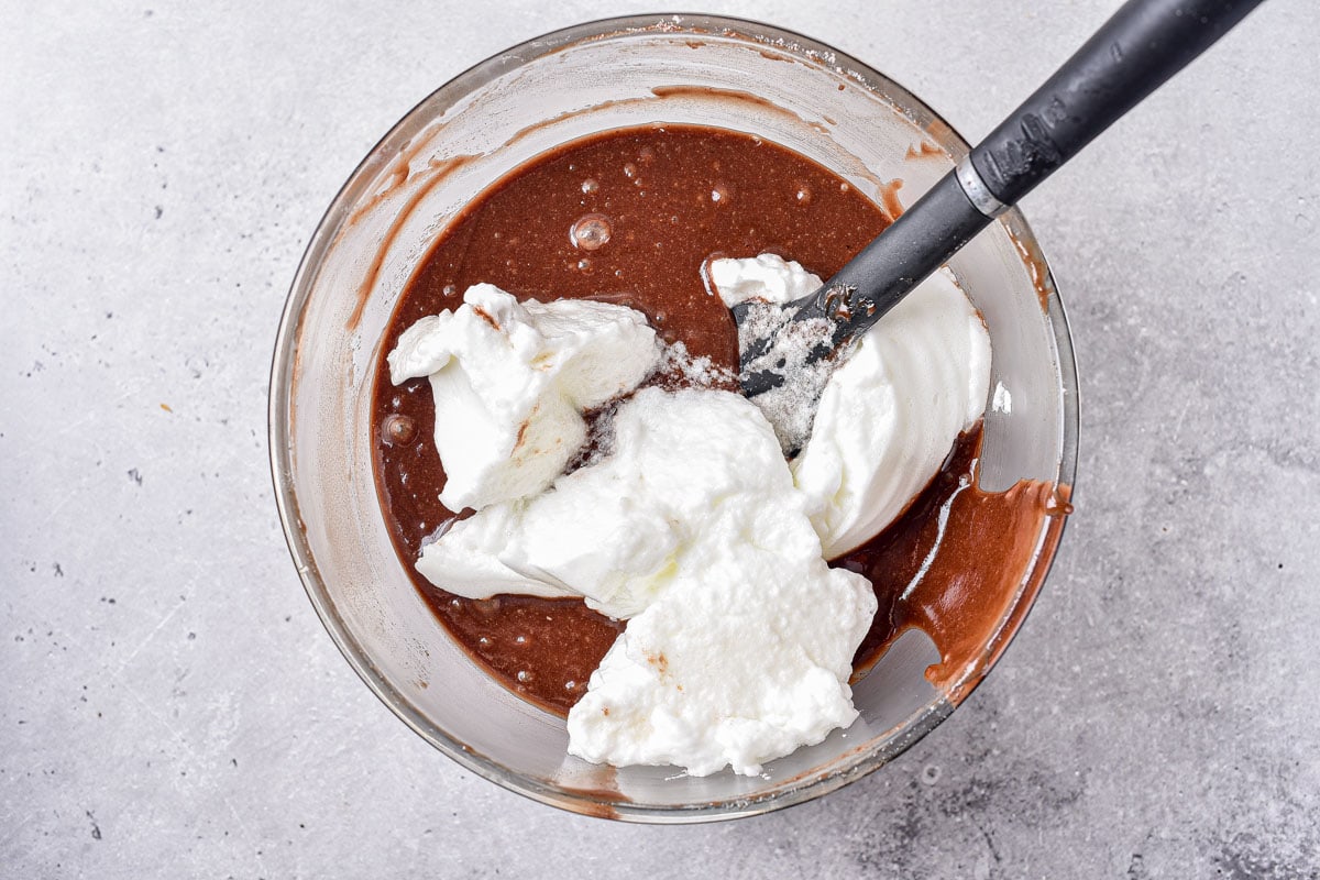 black spatula mixing cream into chocolate batter in glass mixing bowl on counter.
