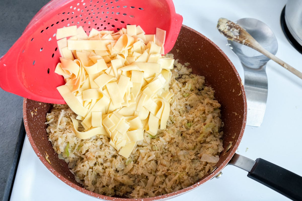 red strainer adding flat egg noodles into pan of fried cabbage.