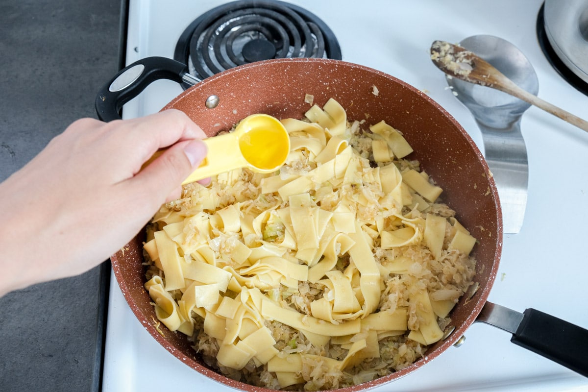 hand adding oil in yellow measuring spoon into pan of fried cabbage.