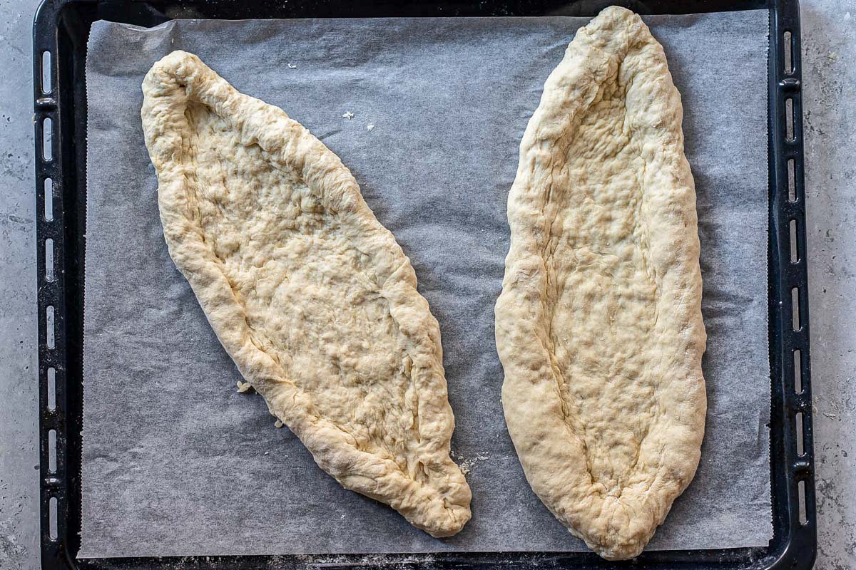 two oval shaped pieces of dough on parchment paper sitting on metal tray.
