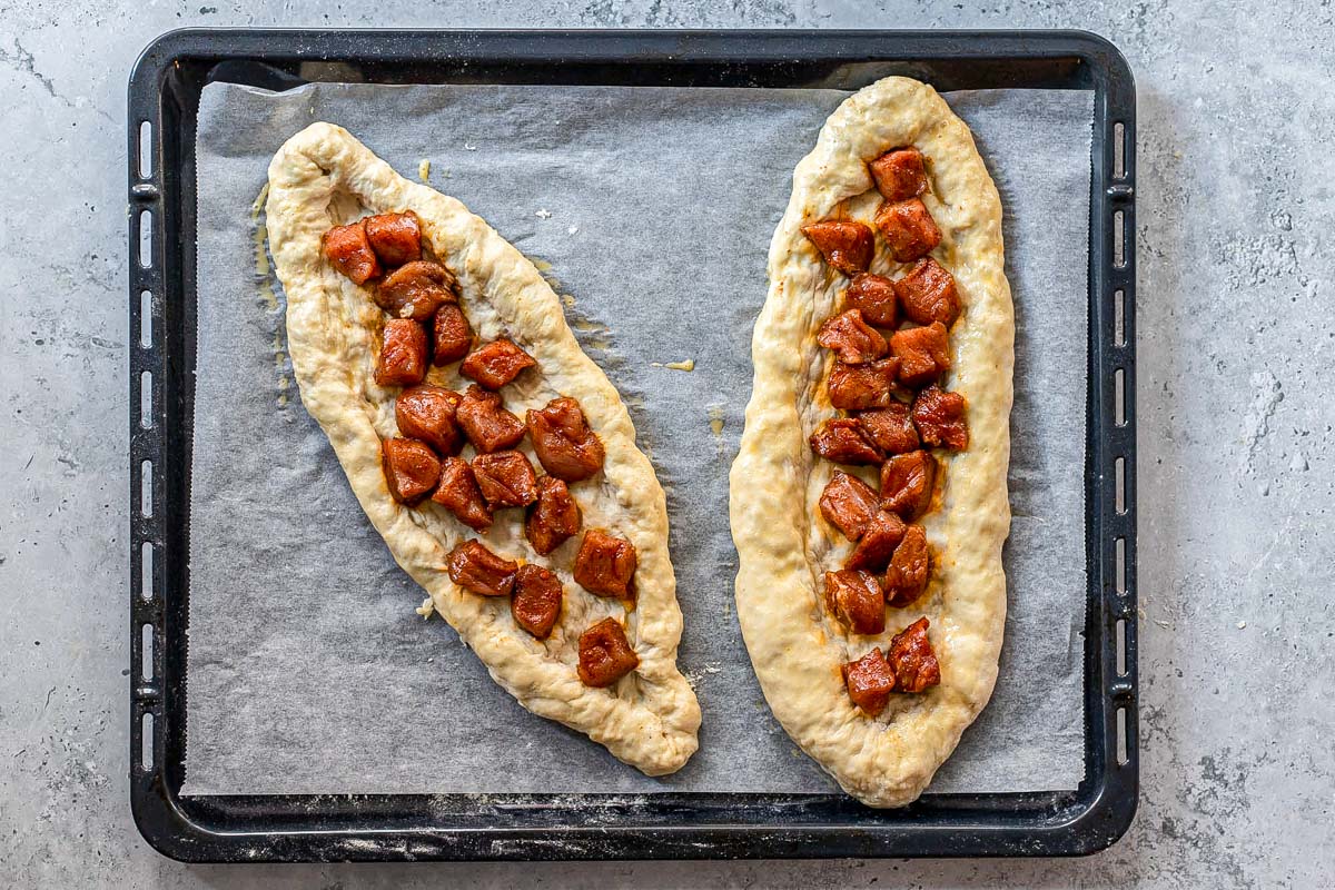raw pork pieces sitting on raw oval shaped dough pizzas on parchment paper.