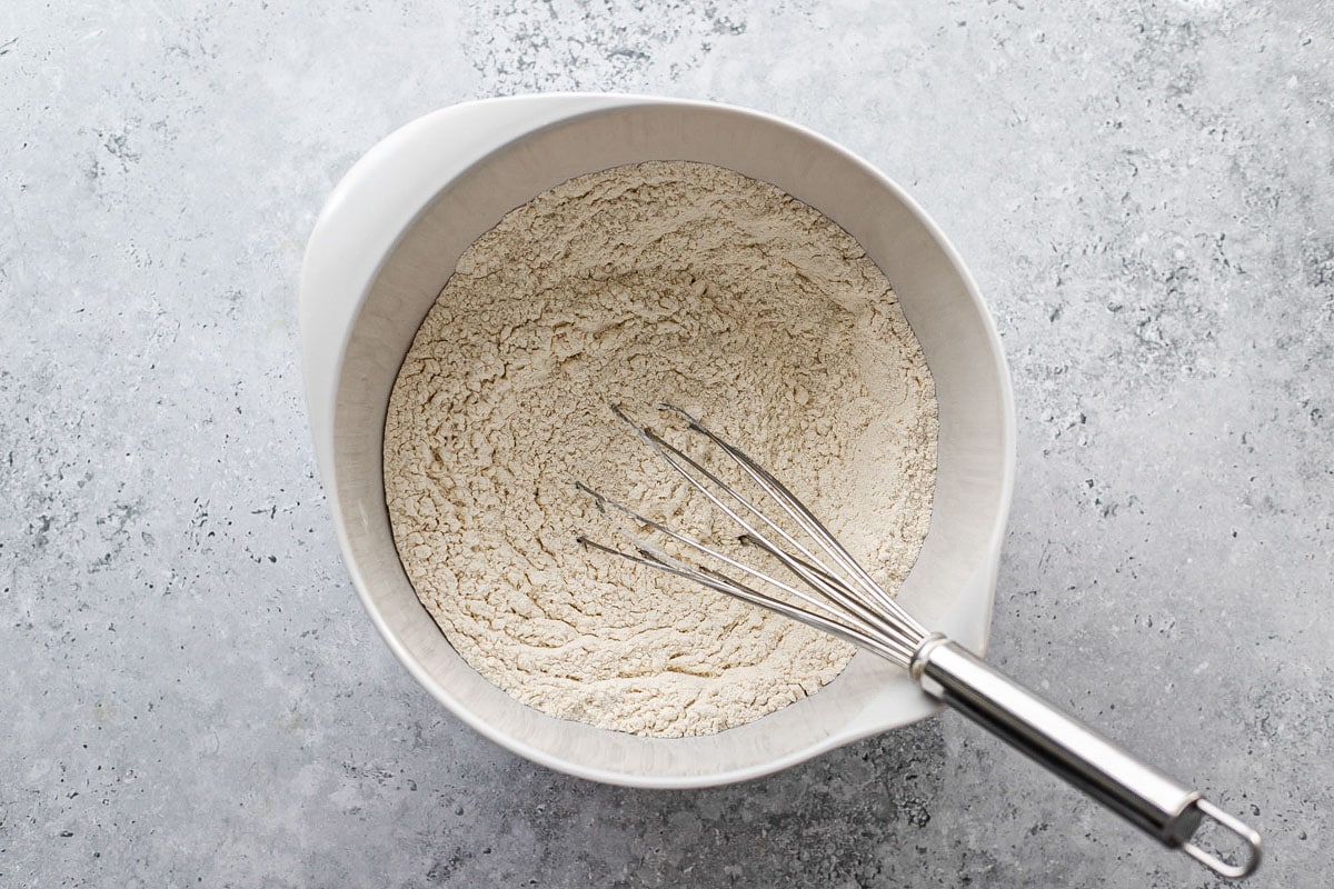 silver whisk mixing dry dough ingredients in white bowl on grey counter.
