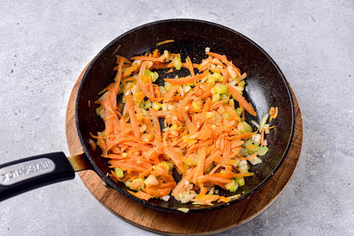 shredded carrots and onions in black frying pan sitting on wooden disc on counter.