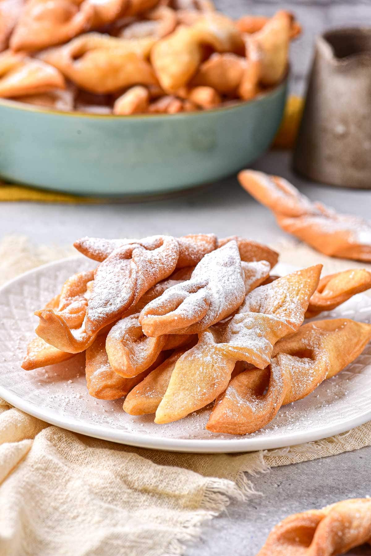crispy fried dough cookies on plate covered in powdered sugar with more behind.