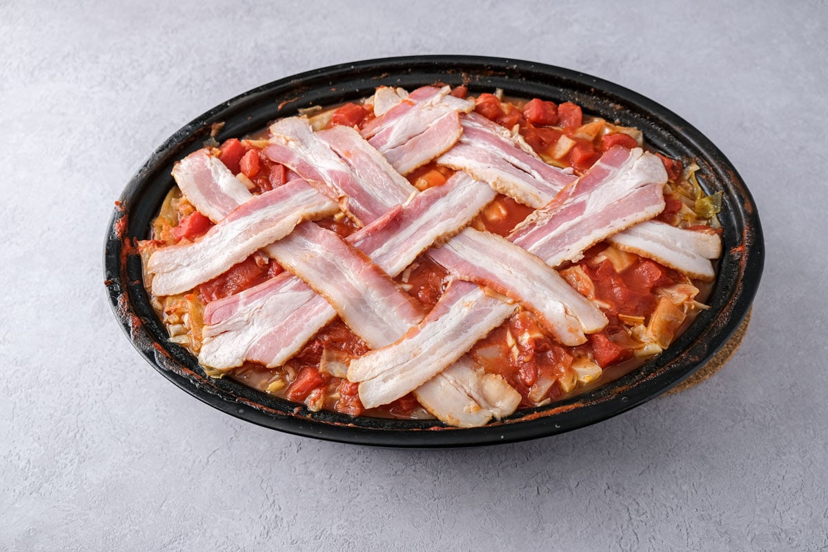 strips of bacon in a lattice on top of red casserole in oval pan.