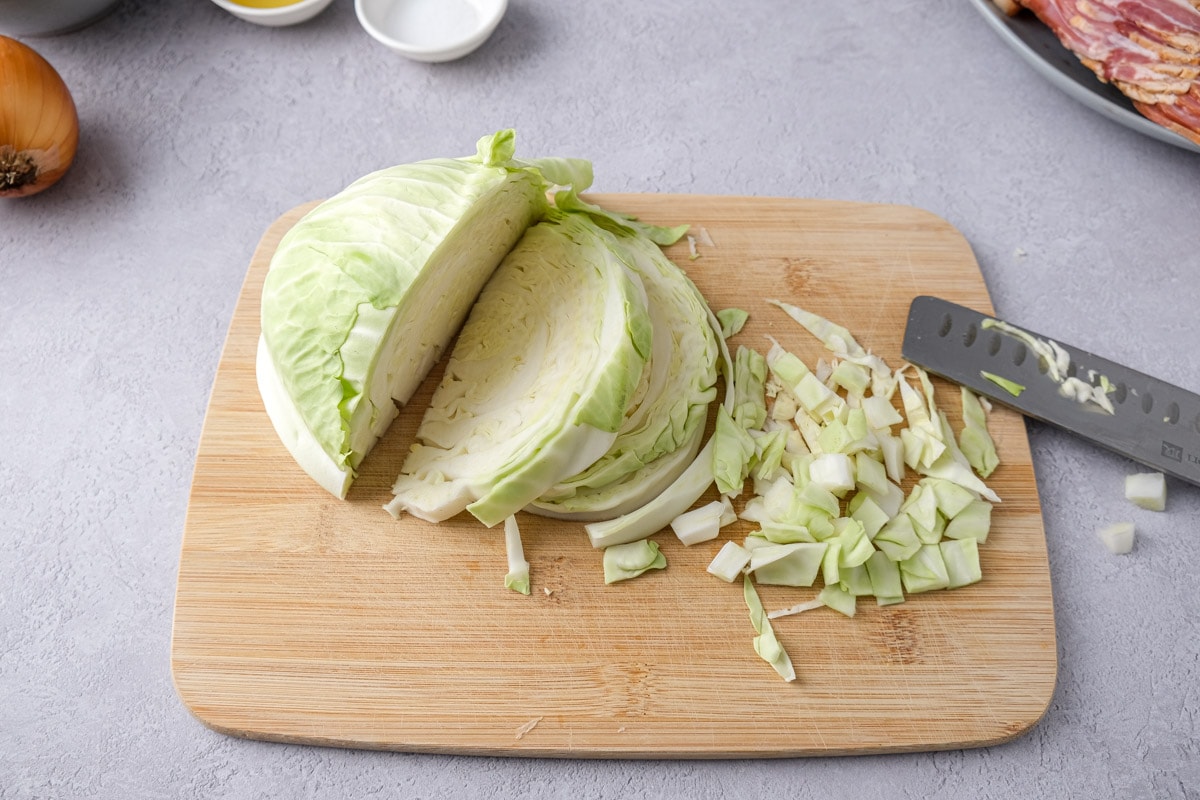 raw cabbage getting chopped on wooden cutting board with knife beside on counter.