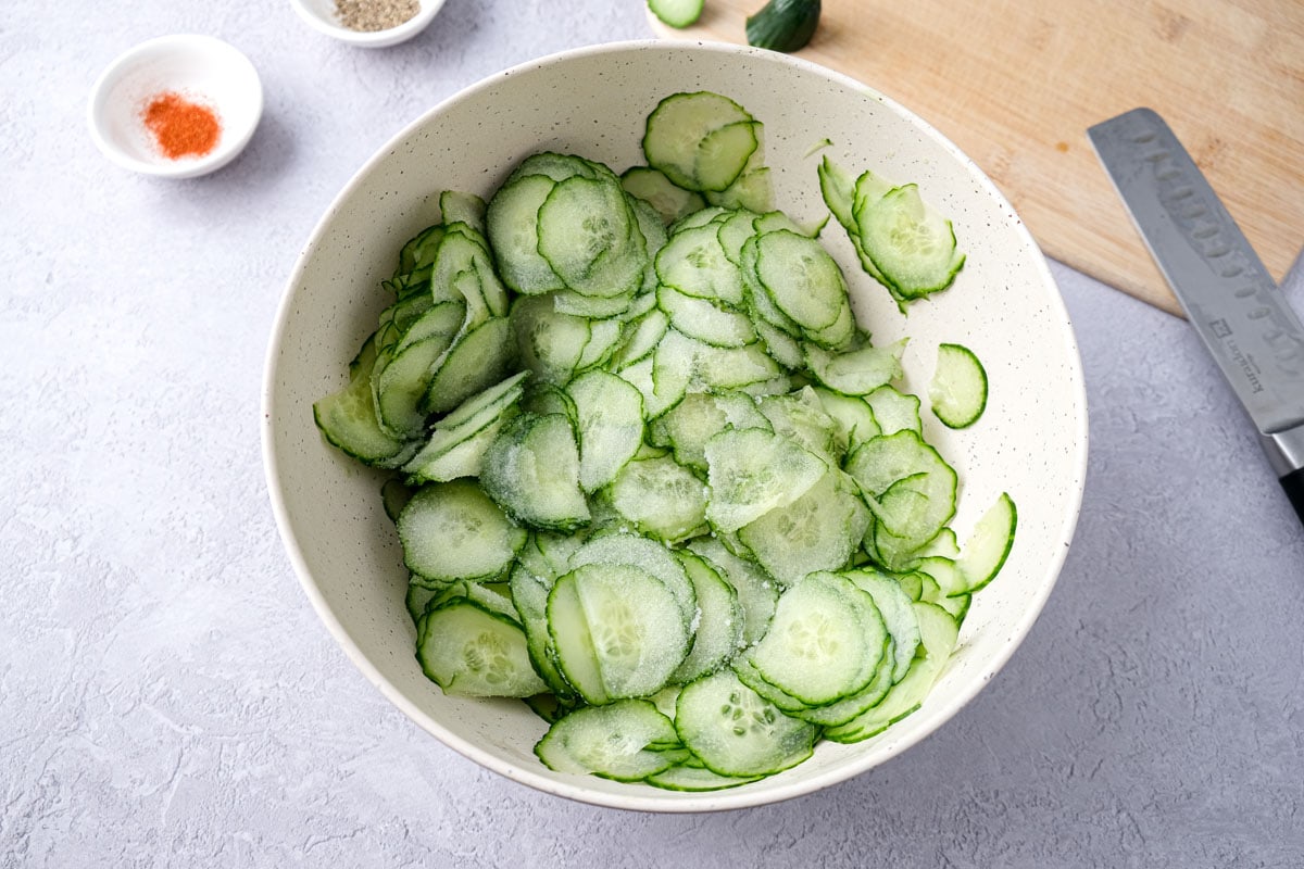 sliced cucumbers in mixing bowl on counter covered in salt.