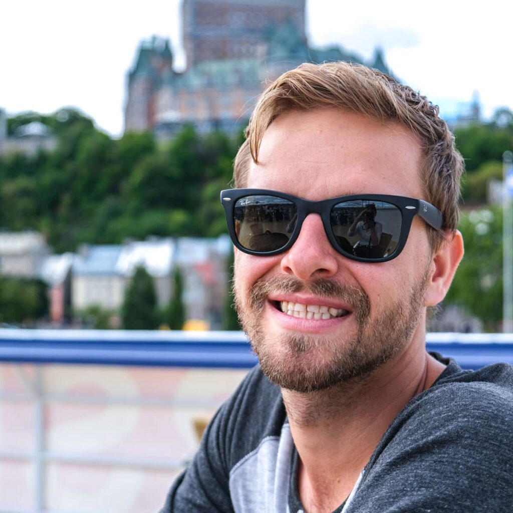 smiling man wearing black sunglasses with old hotel behind.