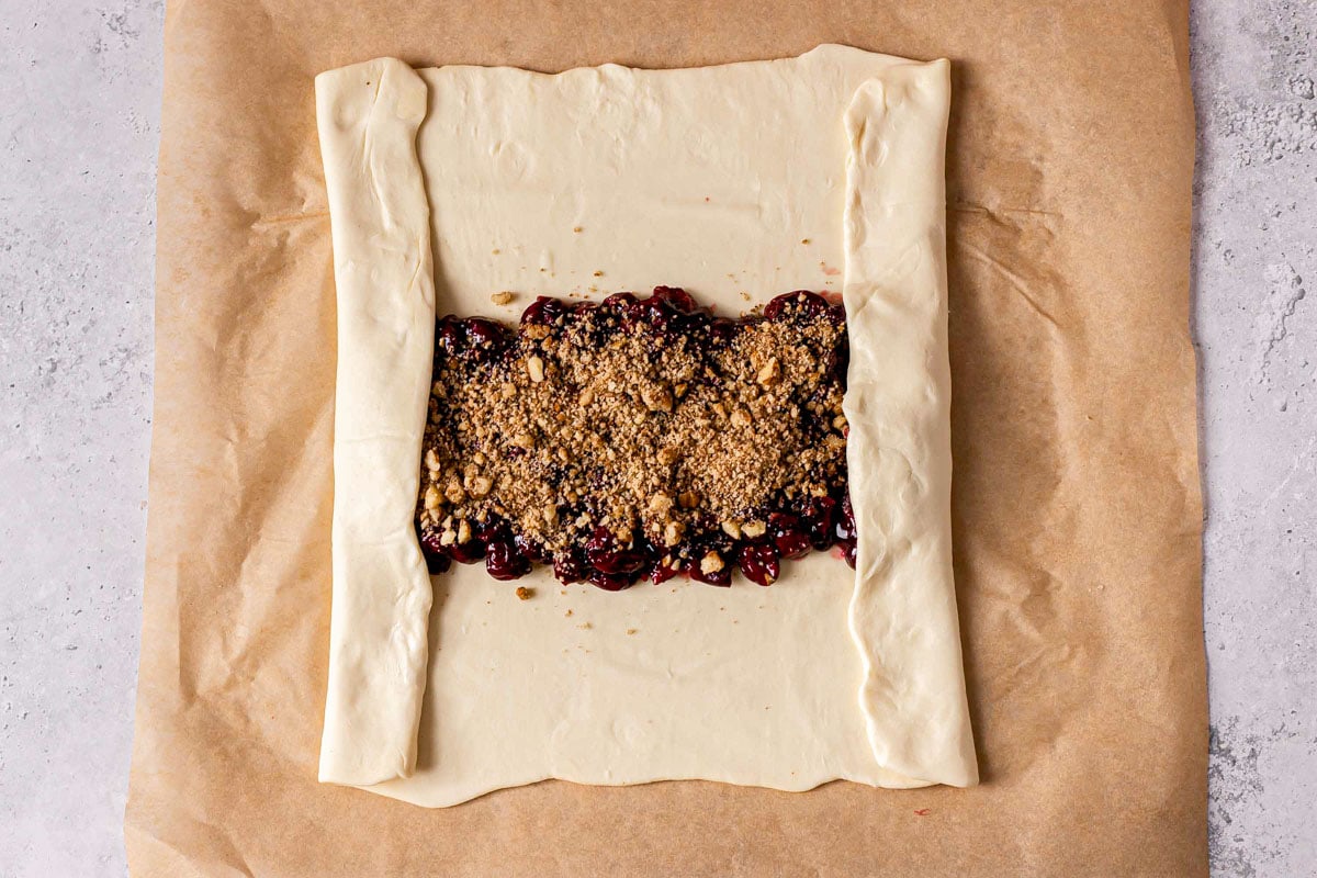 strudel dough filled with cherries and walnuts folded over at both sides sitting on parchment paper.