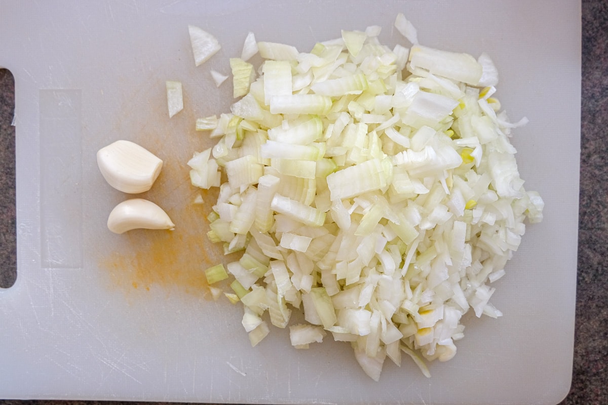 chopped onions and whole garlic on white plastic cutting board.
