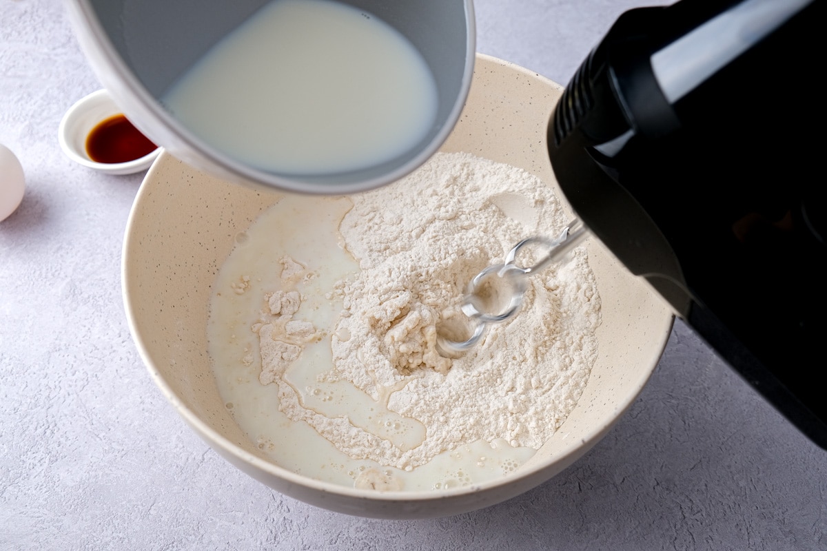 blue bowl pouring milk into mixing bowl filled with dry ingredients with electric mixer beside.