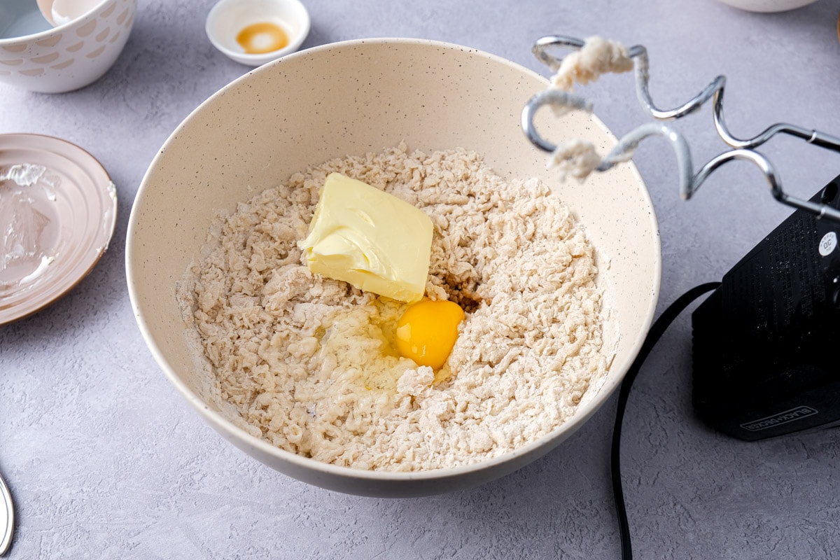 chunk of butter and egg in mixing bowl with raw dough and electric mixer beside.