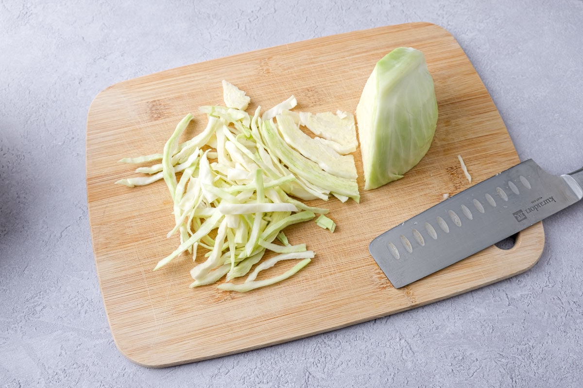 green cabbage on wooden cutting board with knife beside cut into thin strips.