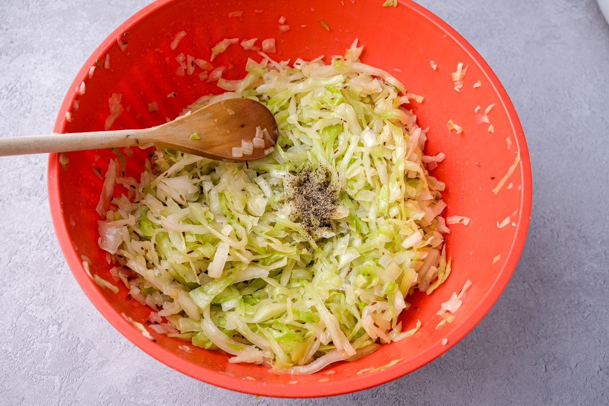 green cabbage in red mixing bowl with ground pepper on top and wooden spoon beside.