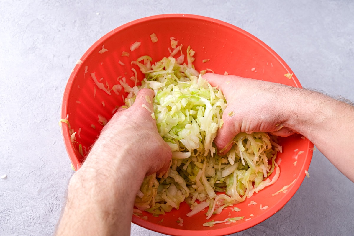 two hands massaging green cabbage in red mixing bowl.