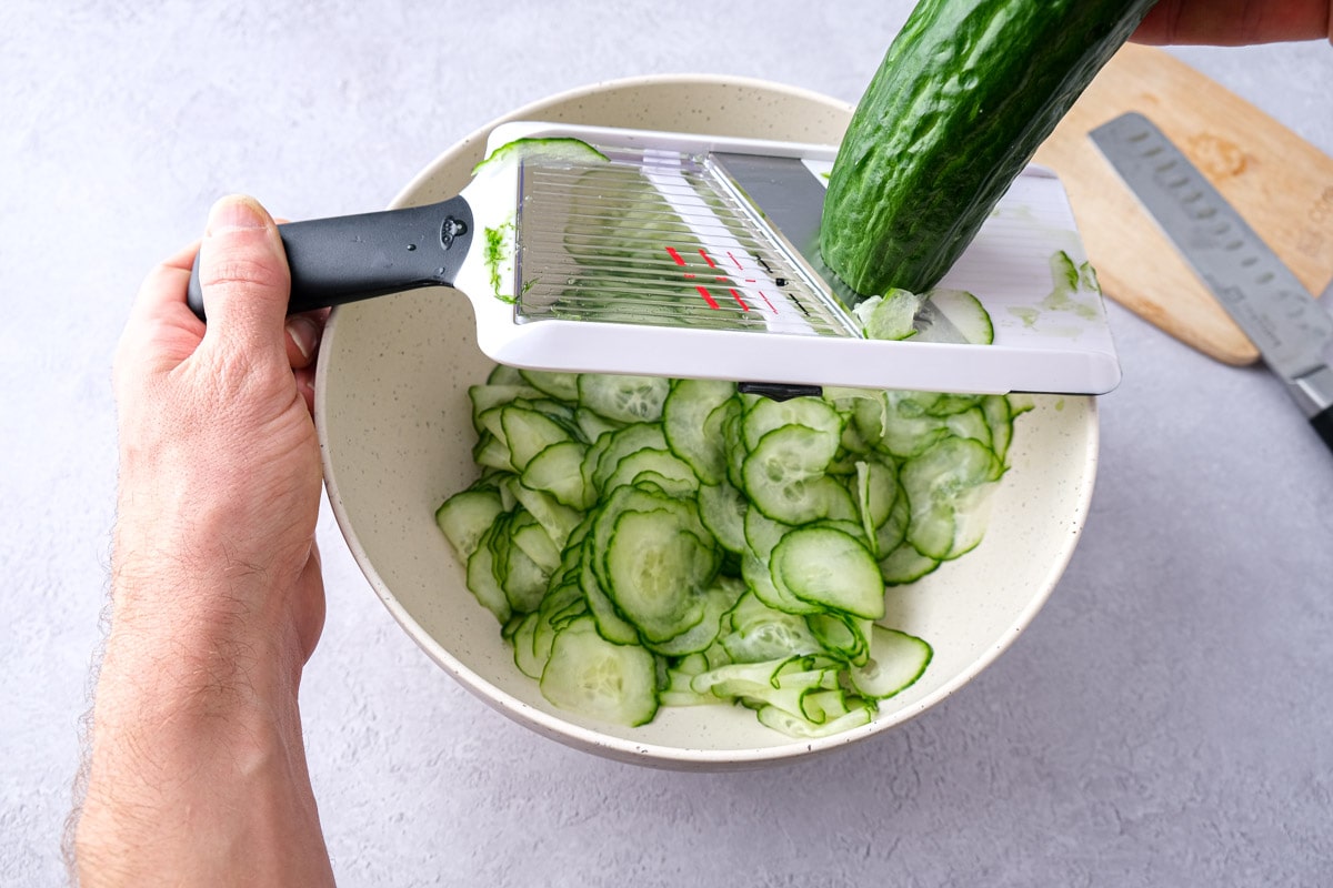 hand holding green cucumber slicing over bowl with mandolin slicer.