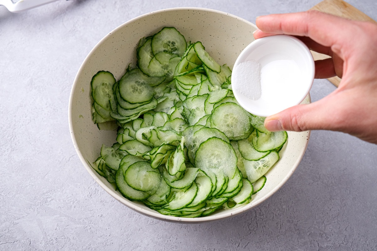 hand adding salt in small bowl to larger bowl of sliced cucumbers.