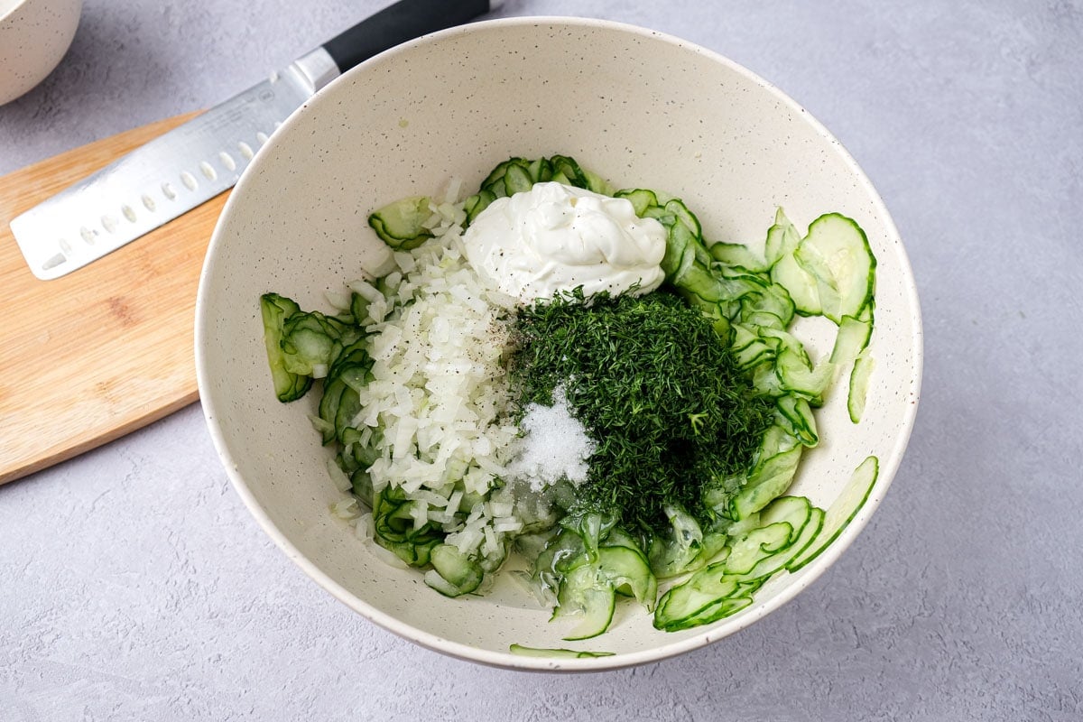 mixing bowl filled with dill, spices, sour cream, and sliced cucumbers.