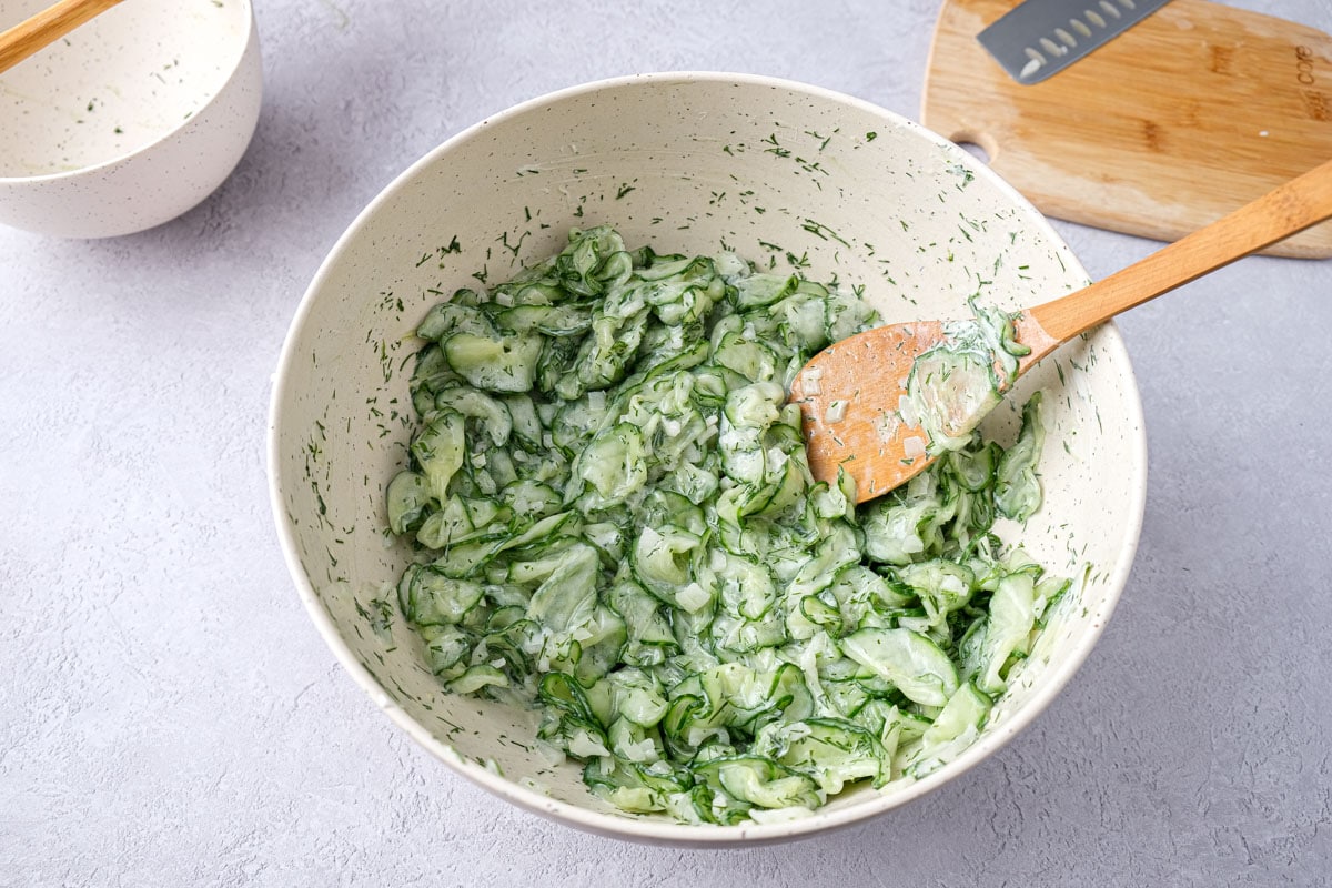 wooden spoon mixing cucumber salad ingredients in white mixing bowl.