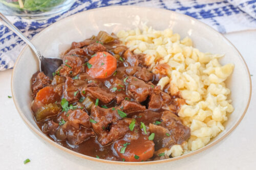 bowl of german beef goulash with spaetzle and fork beside on white counter top.