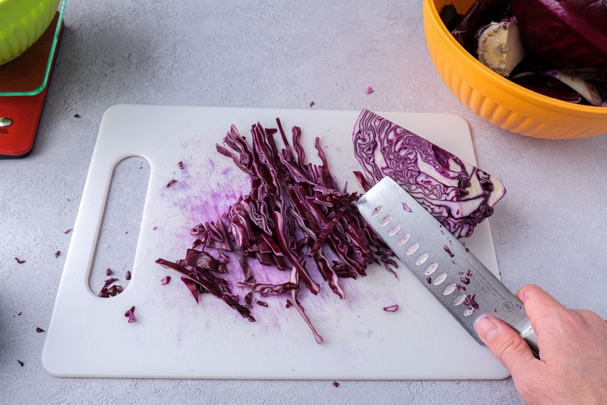 hand holding knife on cutting board beside thinly sliced red cabbage.
