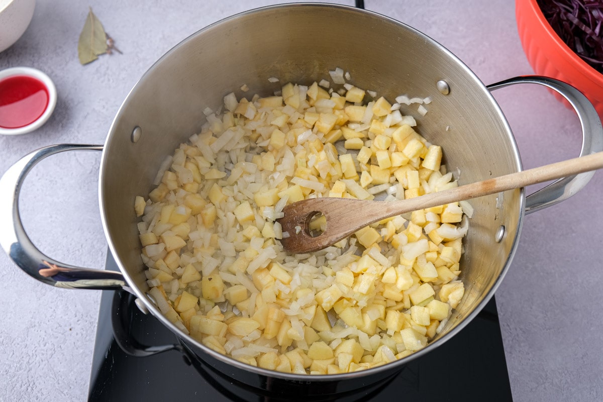 onions and apple frying in large metal pot with wooden spoon stirring around.