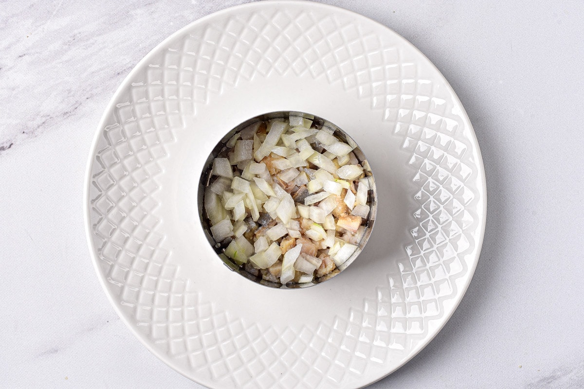 chopped onions on herring pieces in round metal form on white plate.