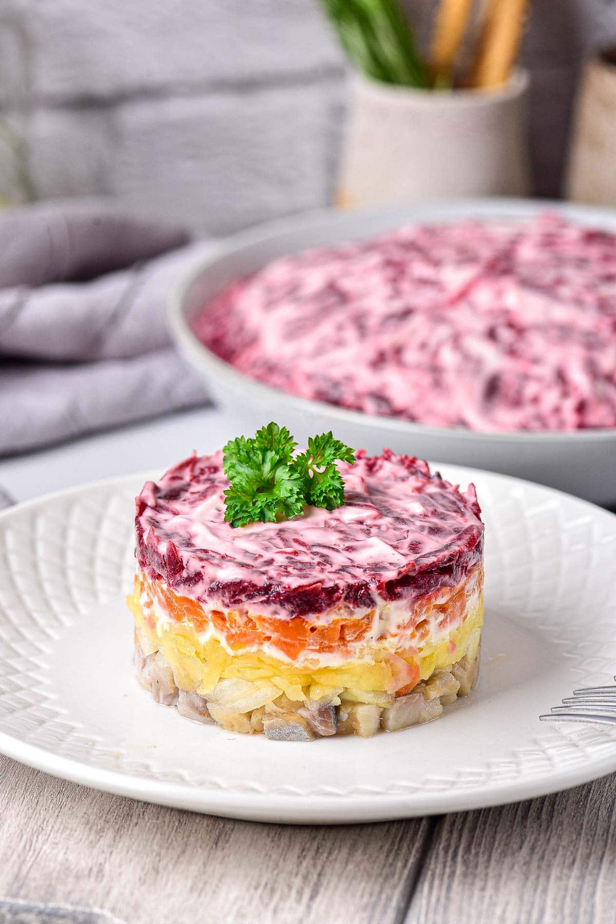 colorful layers of herring salad on white plate with larger salad behind on counter.