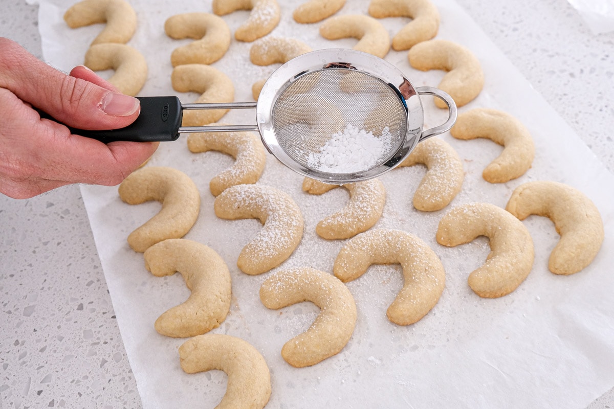 silver strainer sprinkling powdered sugar on crescent cookies in counter.