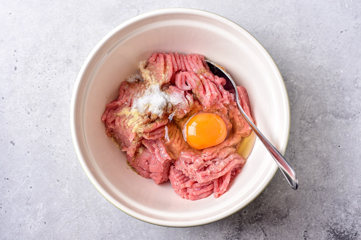 raw egg, meat, and spices in white bowl with silver utensil sticking out.