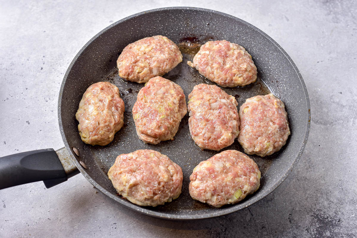 raw meat patties frying in oil in pan with counter around.