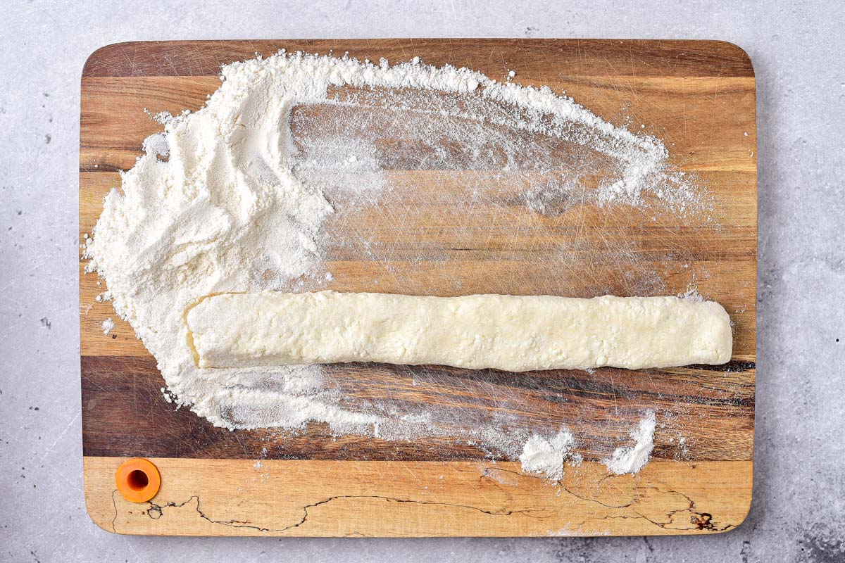 roll of raw dumpling dough on wooden cutting board with flour around.