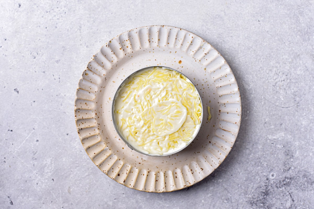 layers of grated cheese and mayo packed into round metal form on plate.