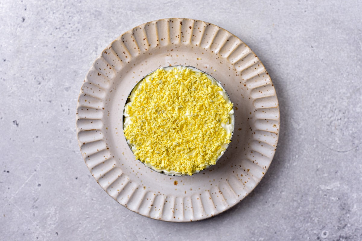 chopped egg yolks on top of layered cold salad in round metal form on plate.
