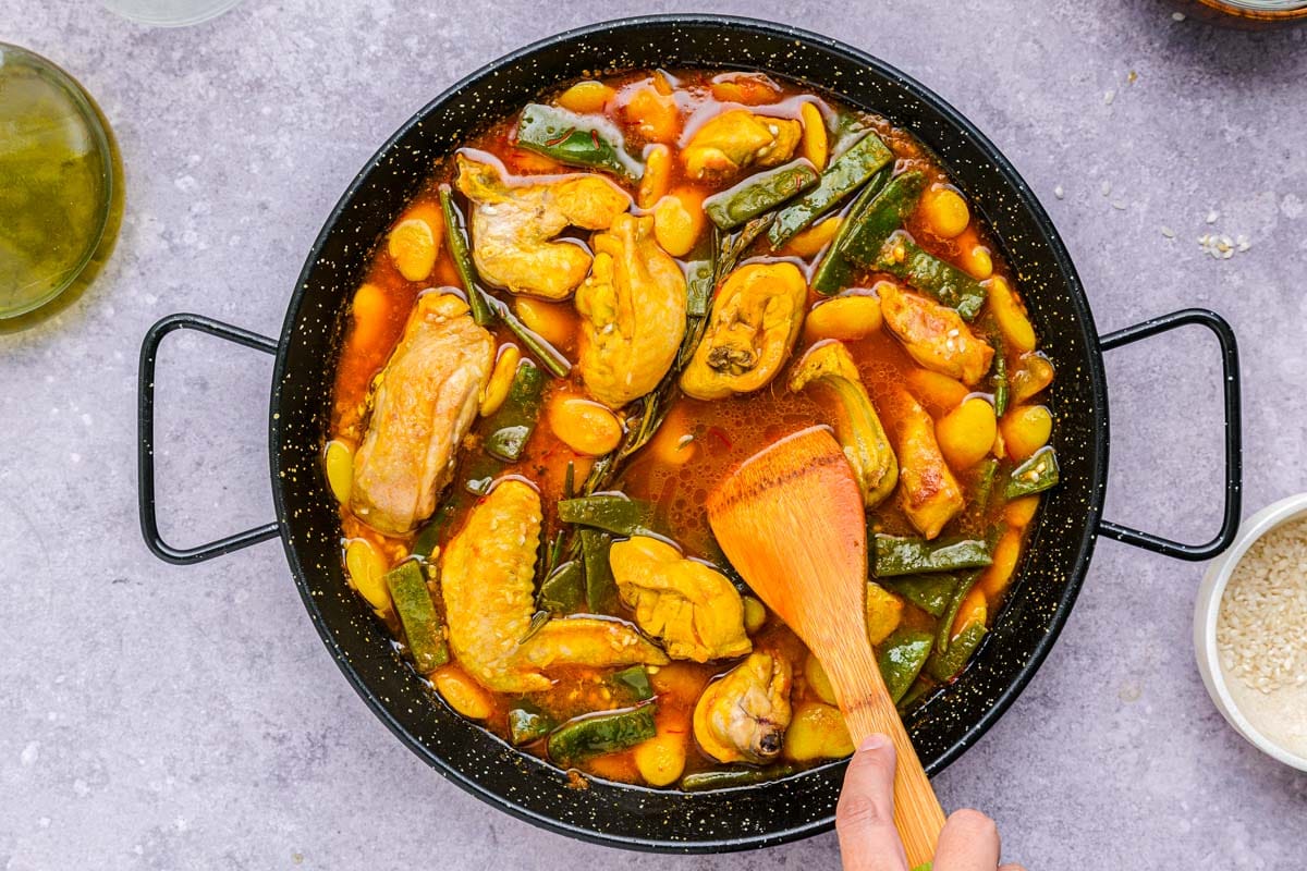 hand stirring paella ingredients cooking in flat black pan.