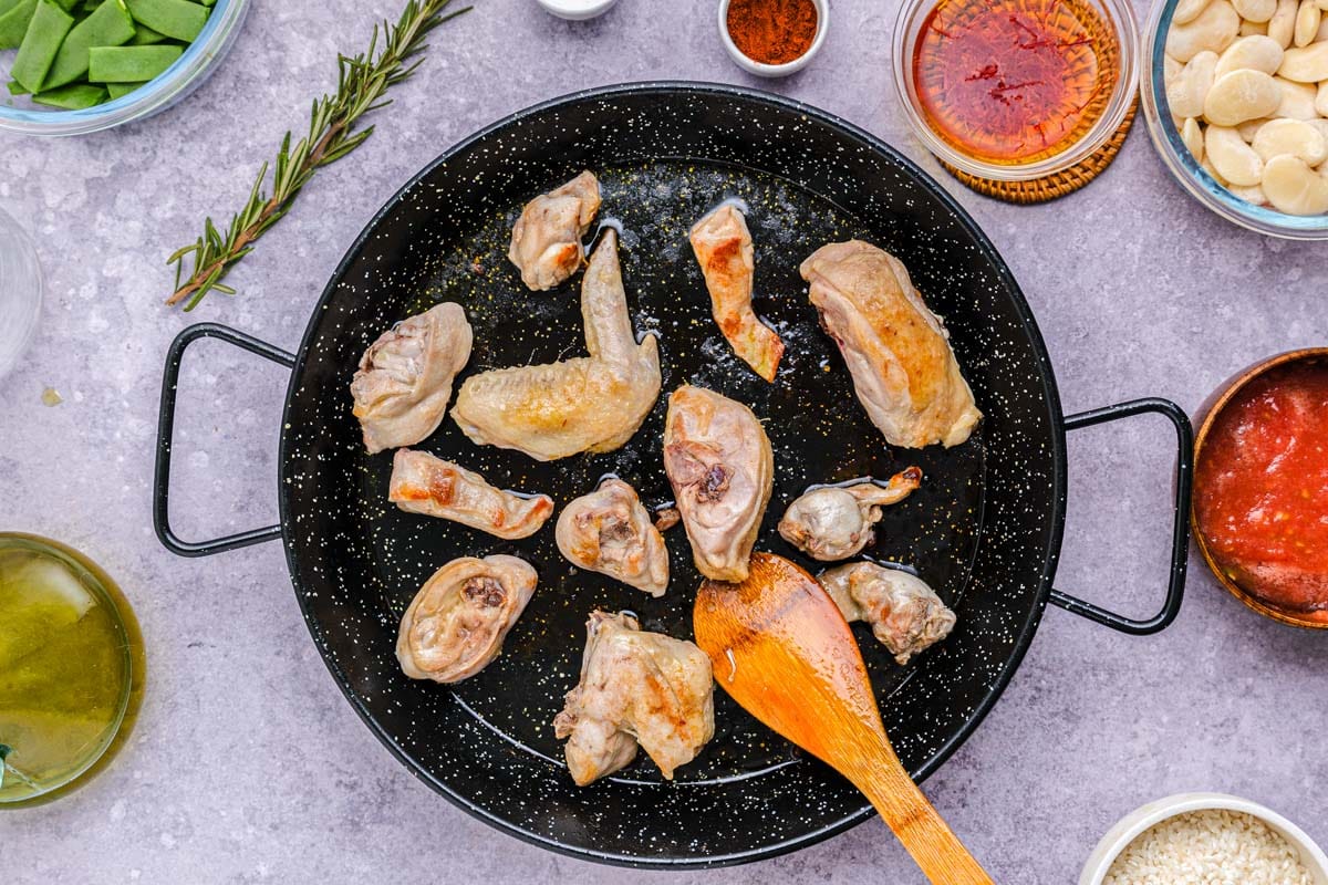 large black pan frying pieces of chicken and rabbit with wooden spoon stirring.