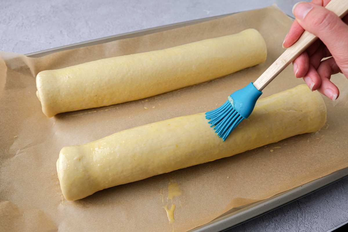 blue brush spreading egg wash on two raw poppy seed rolls on baking pan.