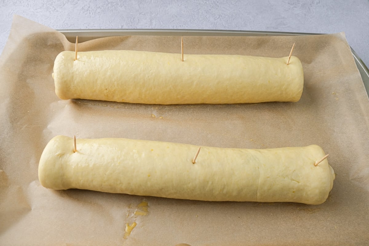 two raw poppy seed rolls with toothpicks sticking out on parchment paper baking tray.