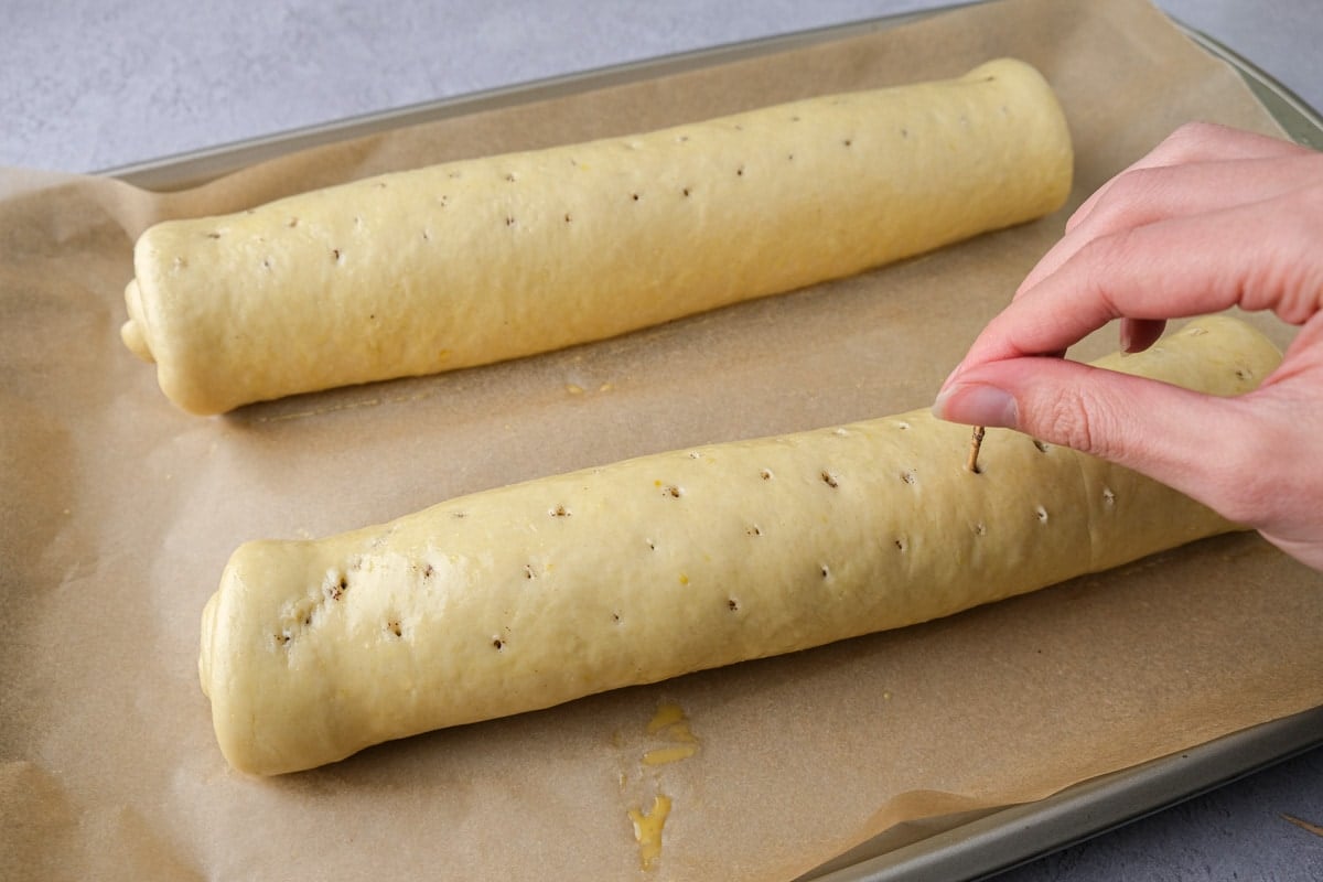 hand holding toothpick piercing holes in top of raw poppy seed rolls.