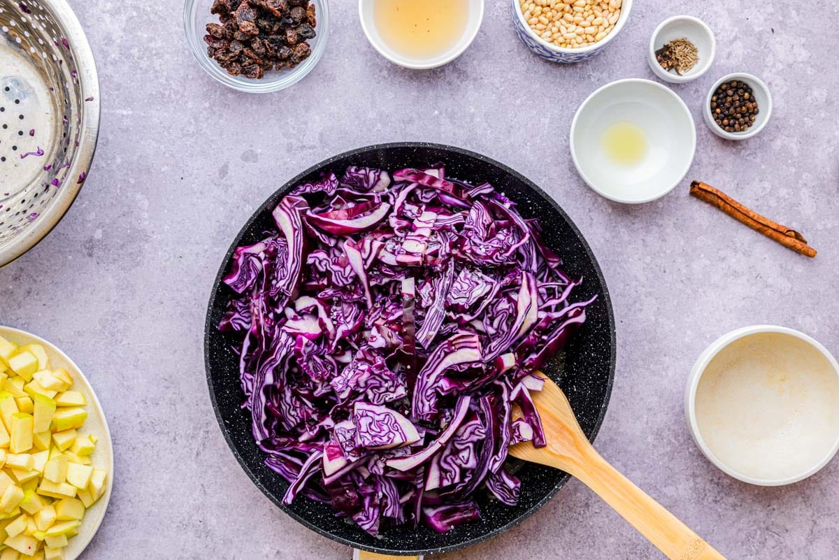 raw red cabbage in black frying pan being stirred with wooden spatula.