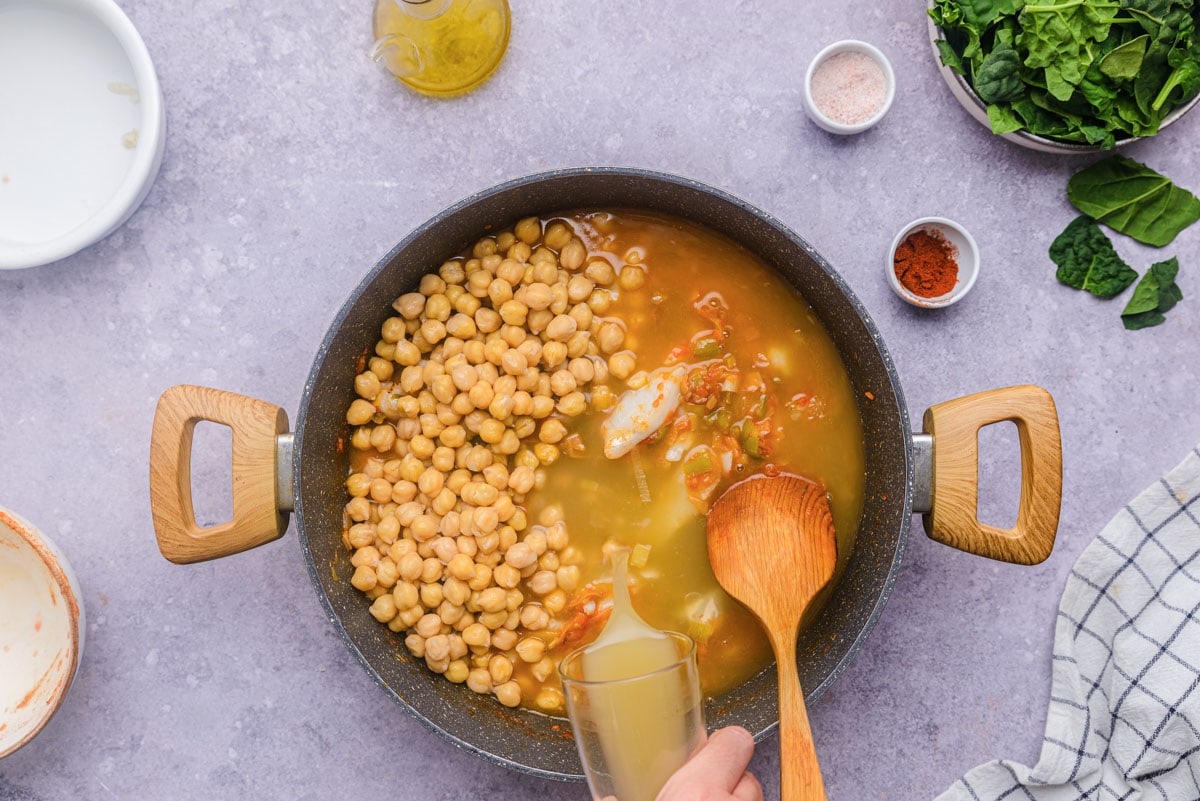 hand pouring broth into large pot of chickpeas and cod on counter.