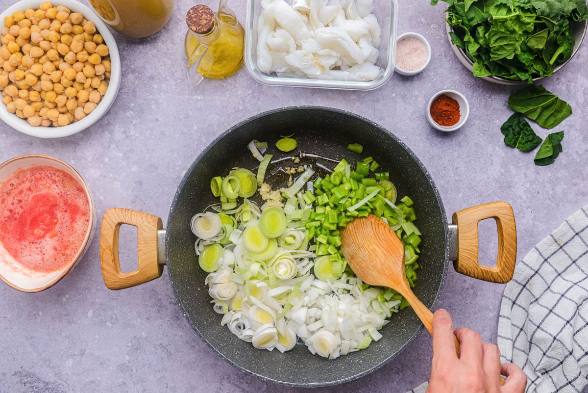 hand holding wooden spatula mixing leeks in black frying pan with ingredients around.