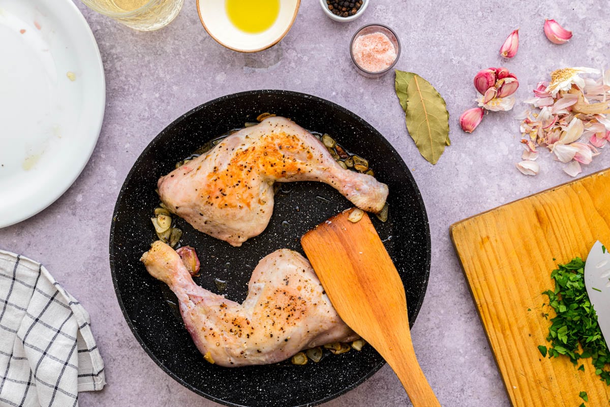 wooden spatula moving cooking chicken legs in black frying pan with other ingredients around.
