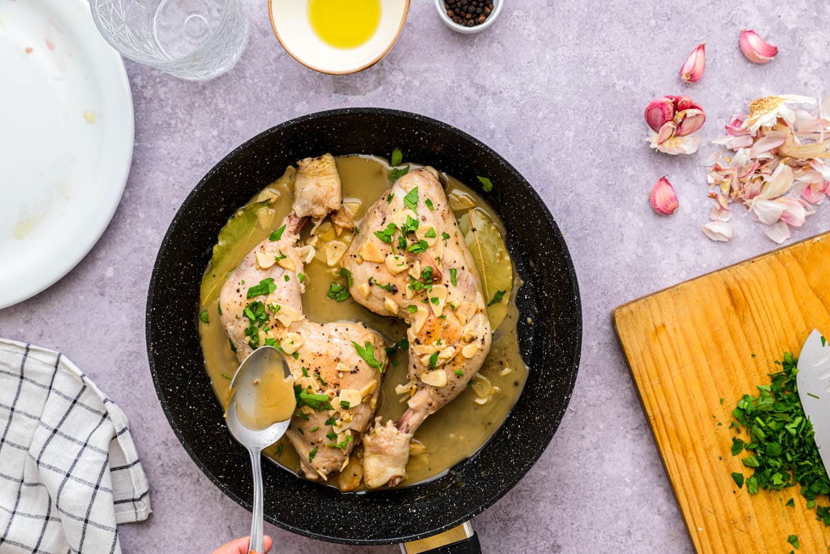 silver spoon pouring thickened garlic sauce over cooking chicken in black pan.