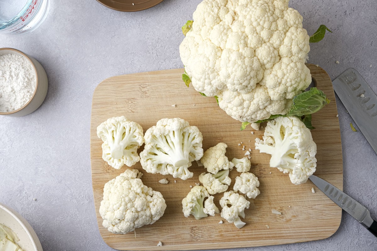 cauliflower florets on wooden cutting board beside larger head of cauliflower.
