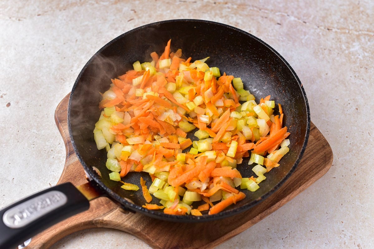 black frying pan full of onions celery and carrot sitting on wooden board on counter.