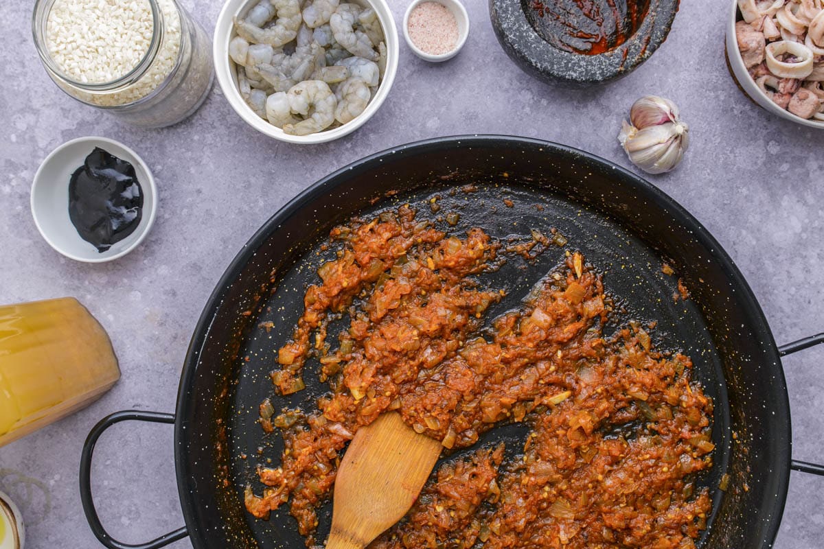 red mixture in flat black frying pan being stirred with wooden spatula.